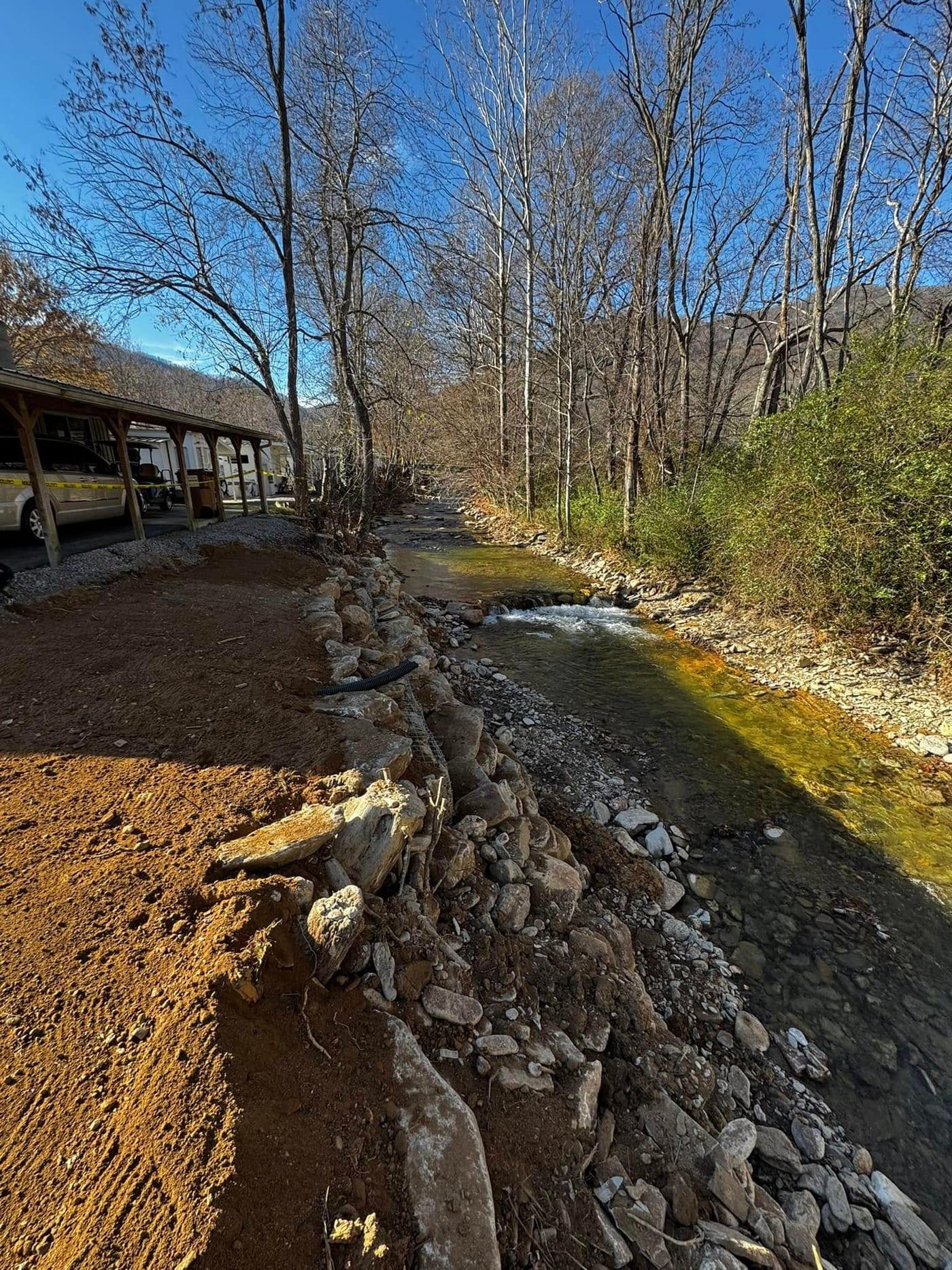 After: the same creek bank rebuilt with a stone-armored toe, a reshaped and graded soil bank, a new drain line and a restored gravel parking area above it.